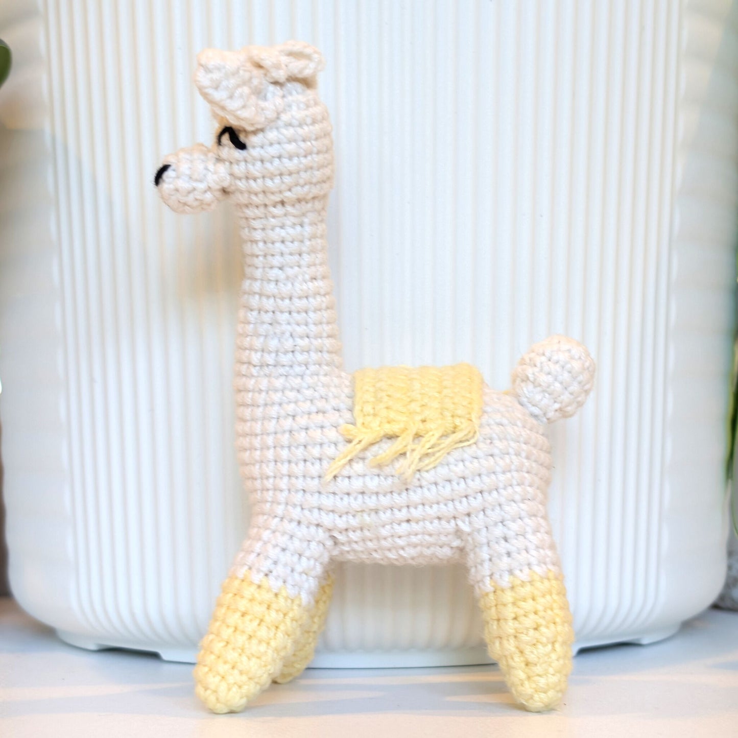 A handmade crocheted llama toy in white with cream yellow accents on its legs, saddle, and ears, standing in front of a white ribbed background.