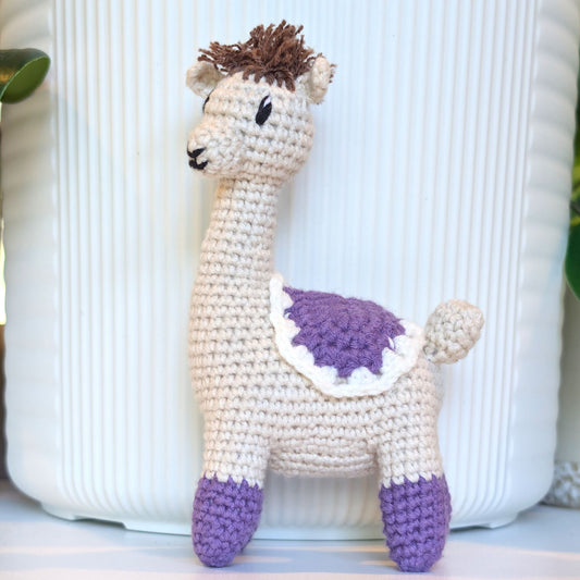A handmade crocheted alpaca toy standing in front of a white ribbed ceramic pot. The toy is cream-colored with purple accents on its belly and feet, and brown hair on its head.