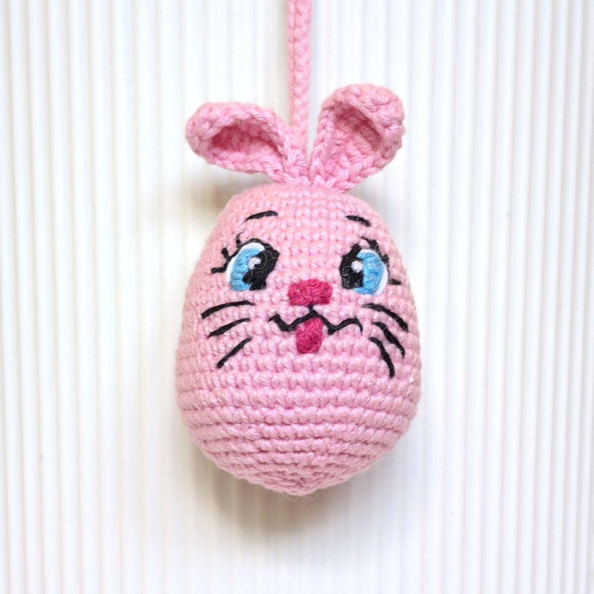 A pink crocheted pink bunny egg ornament with embroidered eyes, nose, whiskers, and upright ears, hanging against a white ribbed background.