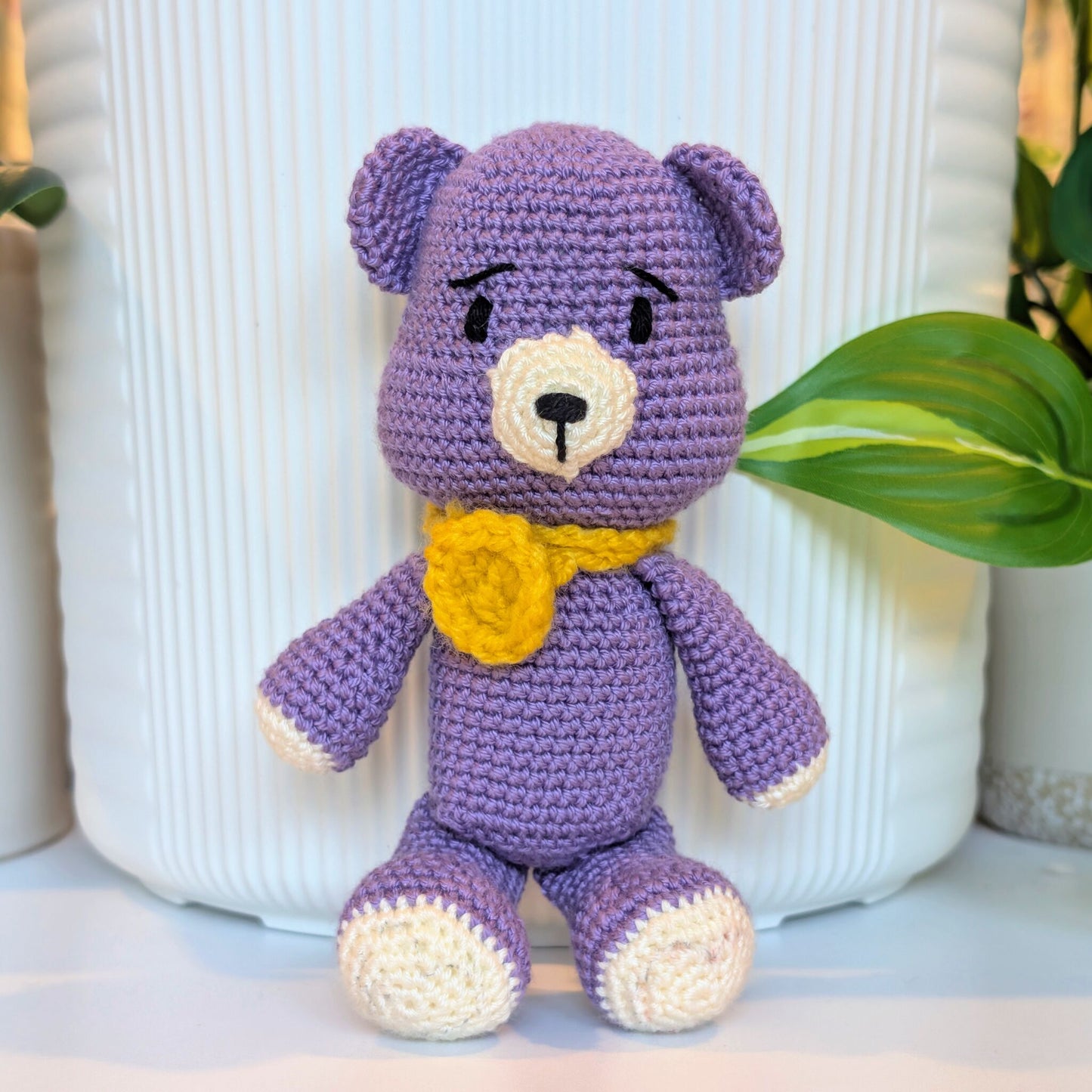 A purple crocheted teddy bear toy with cream-colored paws, snout, and ears, and a yellow flower-shaped collar, posed in front of a white ribbed ceramic vase with green leaves in the background.