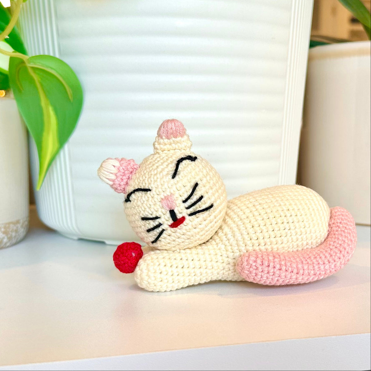 Crocheted cat toy with pink ears and a red ball on a light surface.