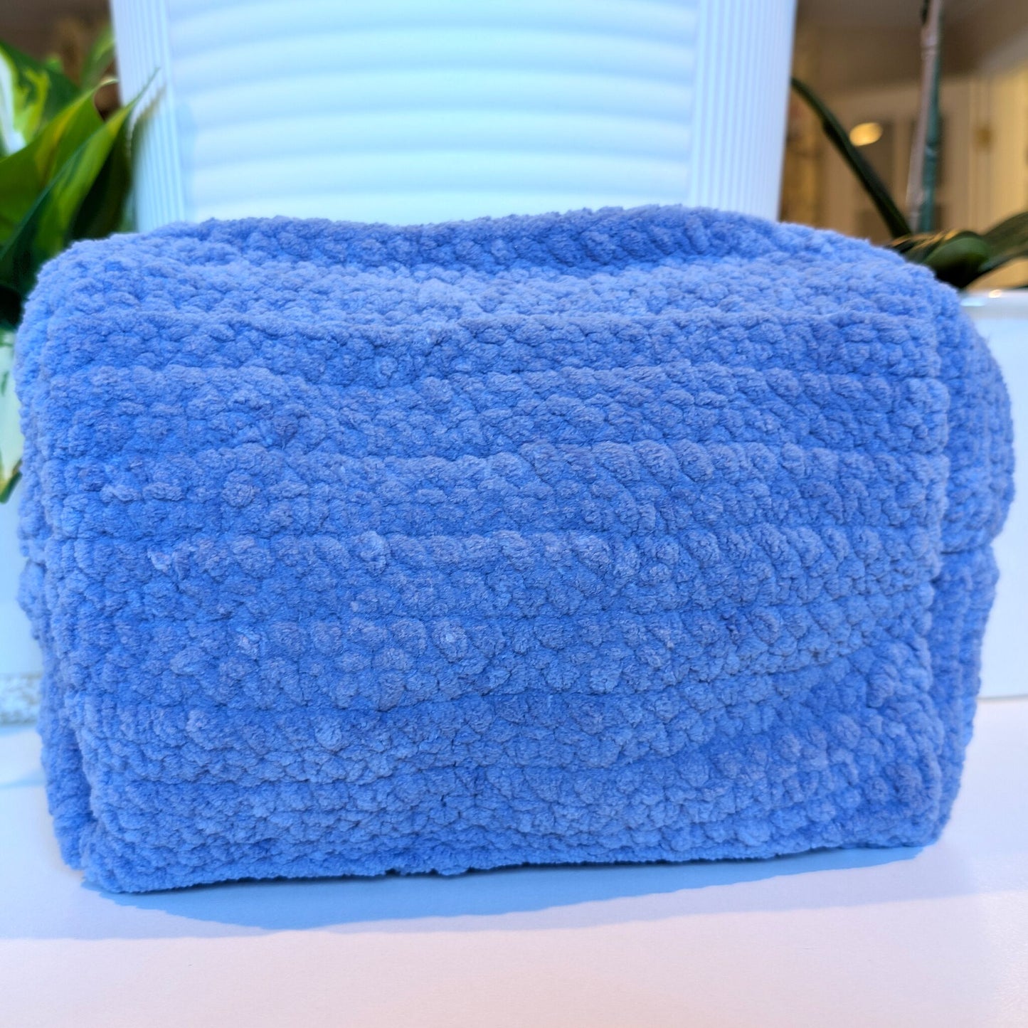 Blue-gray crocheted cosmetic bag on a white surface with a blurred background