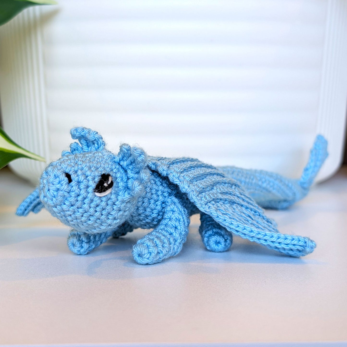 A light blue crocheted dragon toy with large black eyes, resting on a light beige surface in front of a white ribbed background.