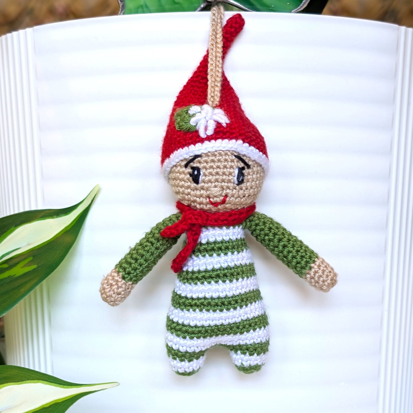 Crocheted elf ornament with a red hat and moss green striped outfit with an edelweis flower in its hat on a white surface.