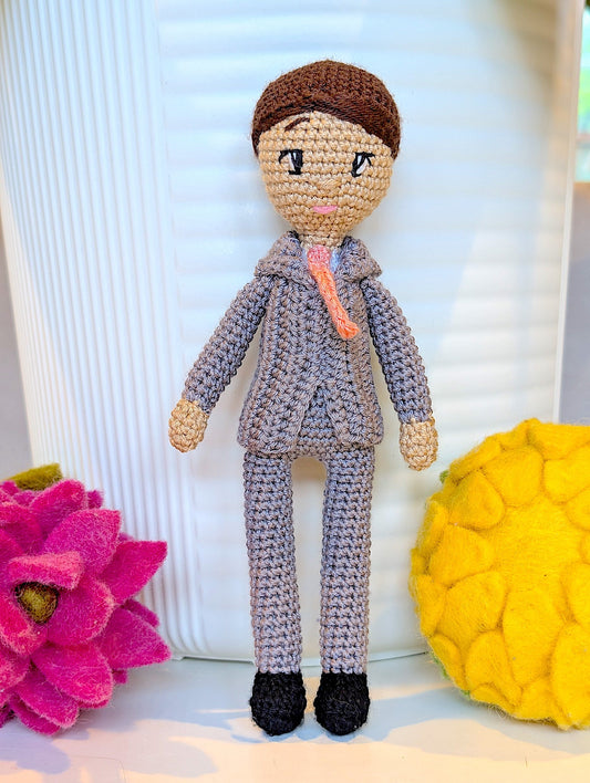 Crocheted boy doll in a grey suit with a pink bow tie and black shoes