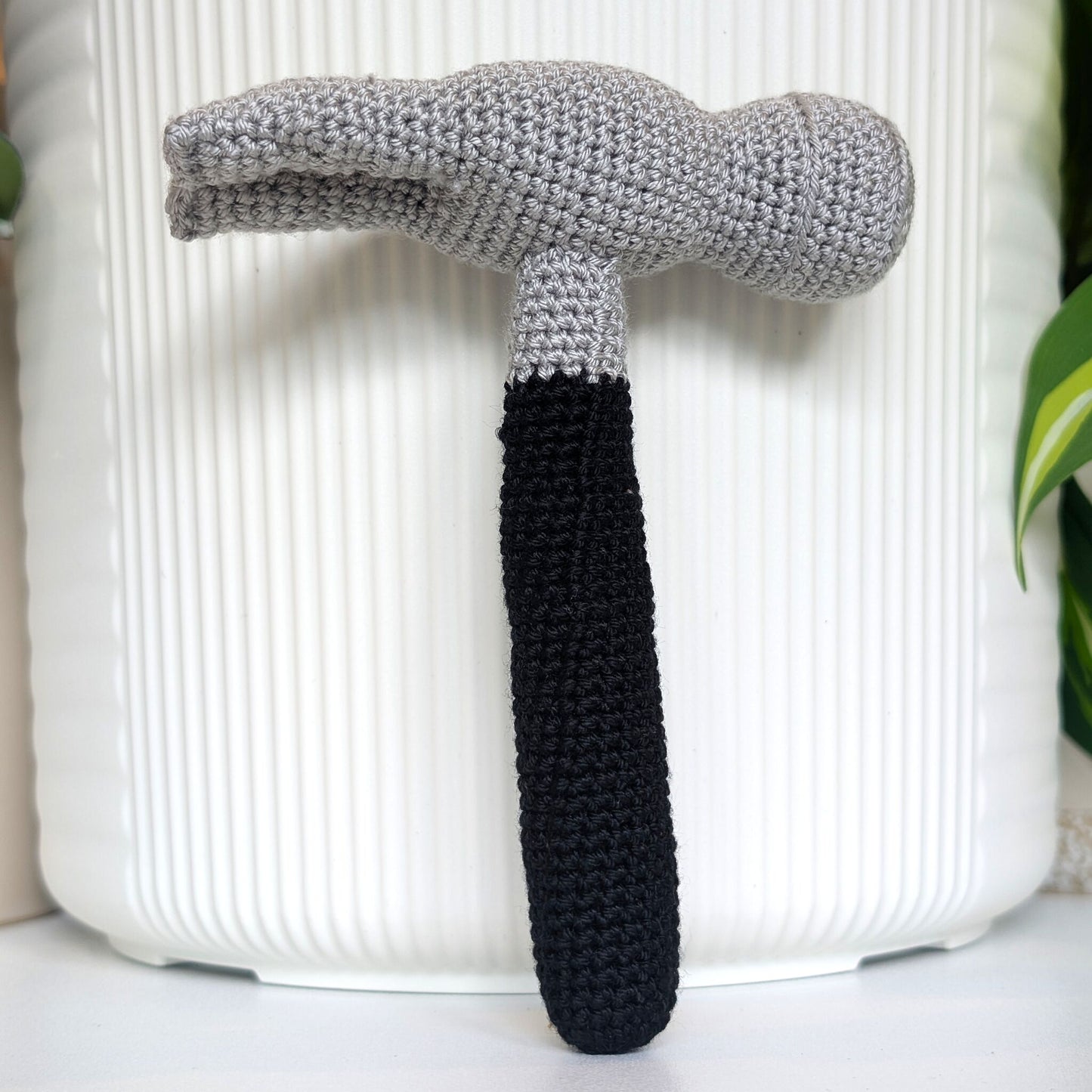 A crocheted hammer with a light gray head and a black handle, standing upright in front of a white ribbed cylindrical planter pot.