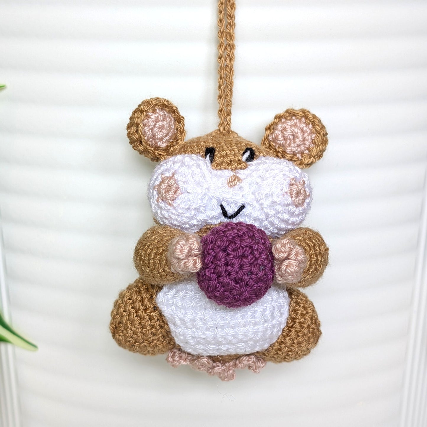 Crochet sand brown hamster bag charm with a string holding a plum colored ball