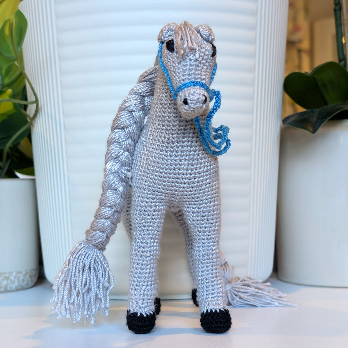 A crocheted plush horse toy with a bright gray body, white mane and tail, black hooves, and a bright blue bridle, standing in front of a white ribbed vase with potted plants in the background.