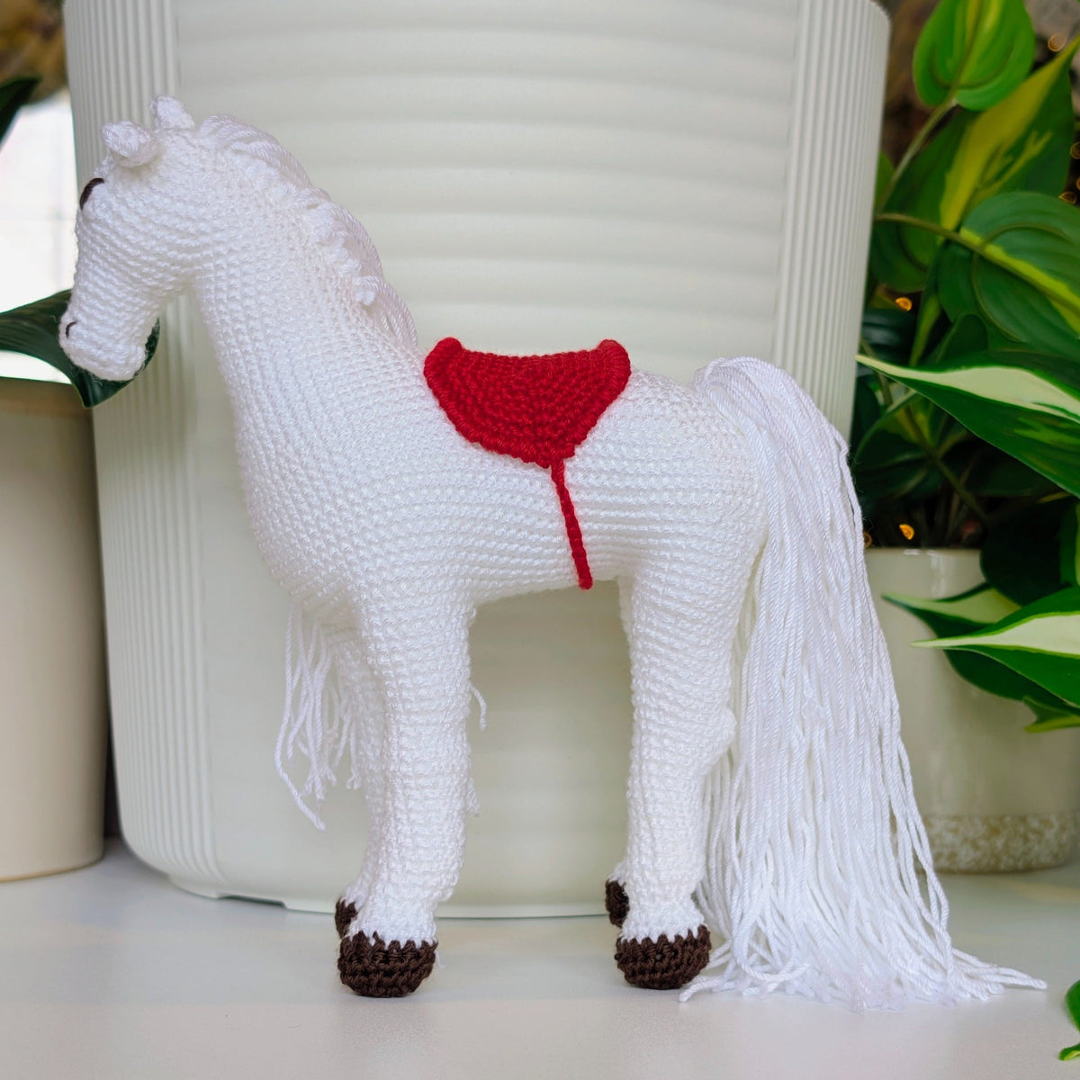White crochet horse with a red saddle on a light surface with plants in the background