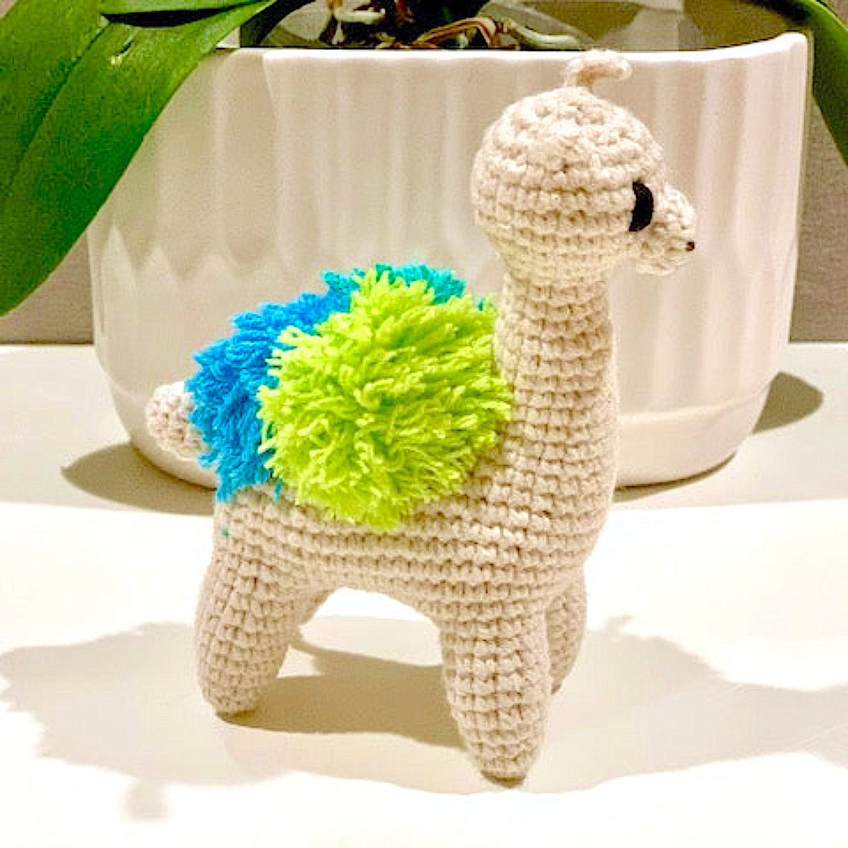 Crocheted llama toy with a blue and green accents on a white surface.