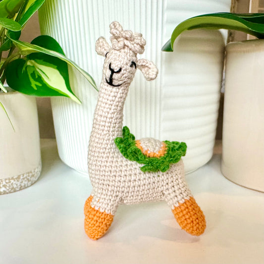 Crocheted llama toy with green leaf detail on a white surface with plants in the background