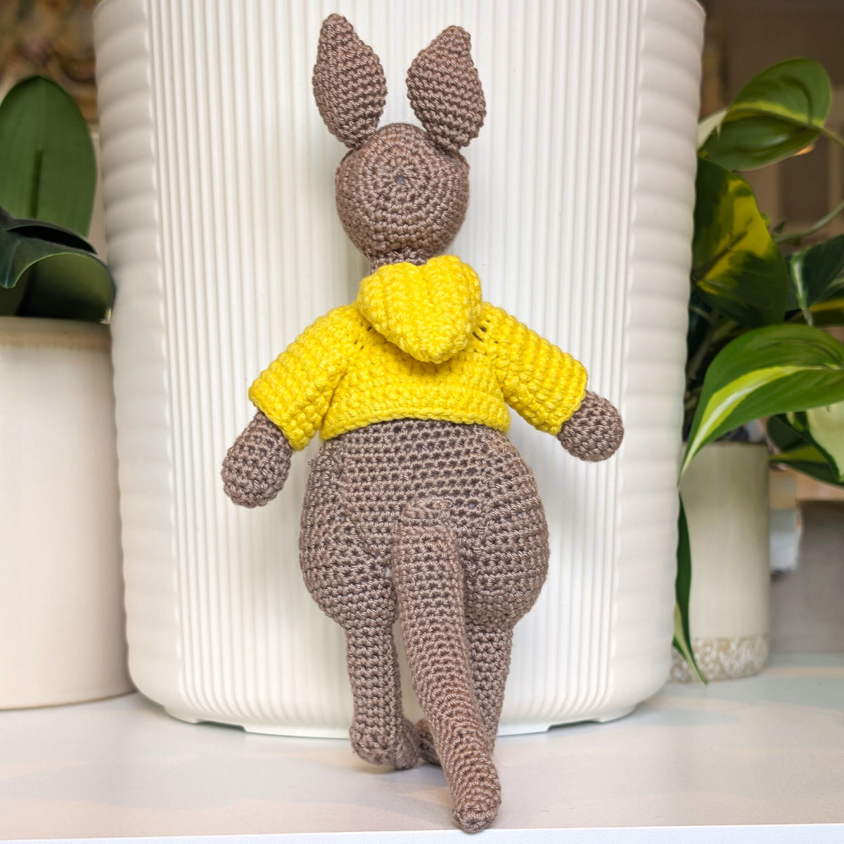 A crocheted kangaroo toy wearing a bright yellow hoodie, positioned in front of a white ribbed ceramic pot with green plants in the background.