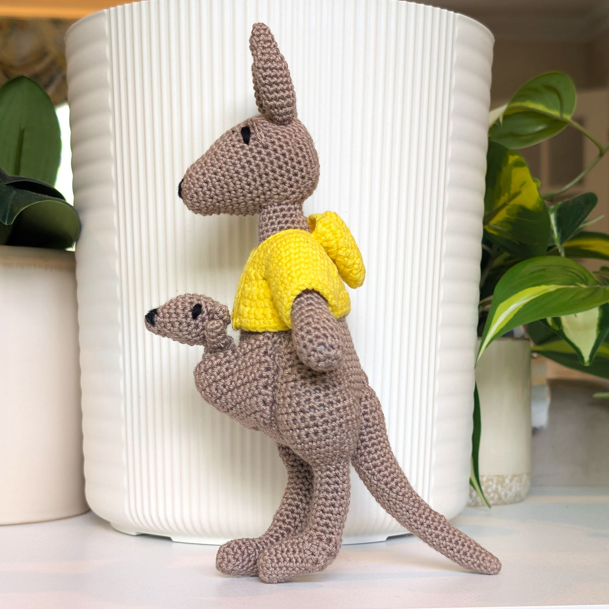 A crocheted kangaroo toy with a joey inside pouch, wearing a bright yellow shirt, posed in front of a white ribbed cylindrical planter with green plants in the background.