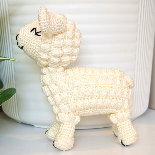 A cream-colored crocheted lamb figurine with black stitched eyes and nose, standing in front of a white ribbed ceramic pot.
