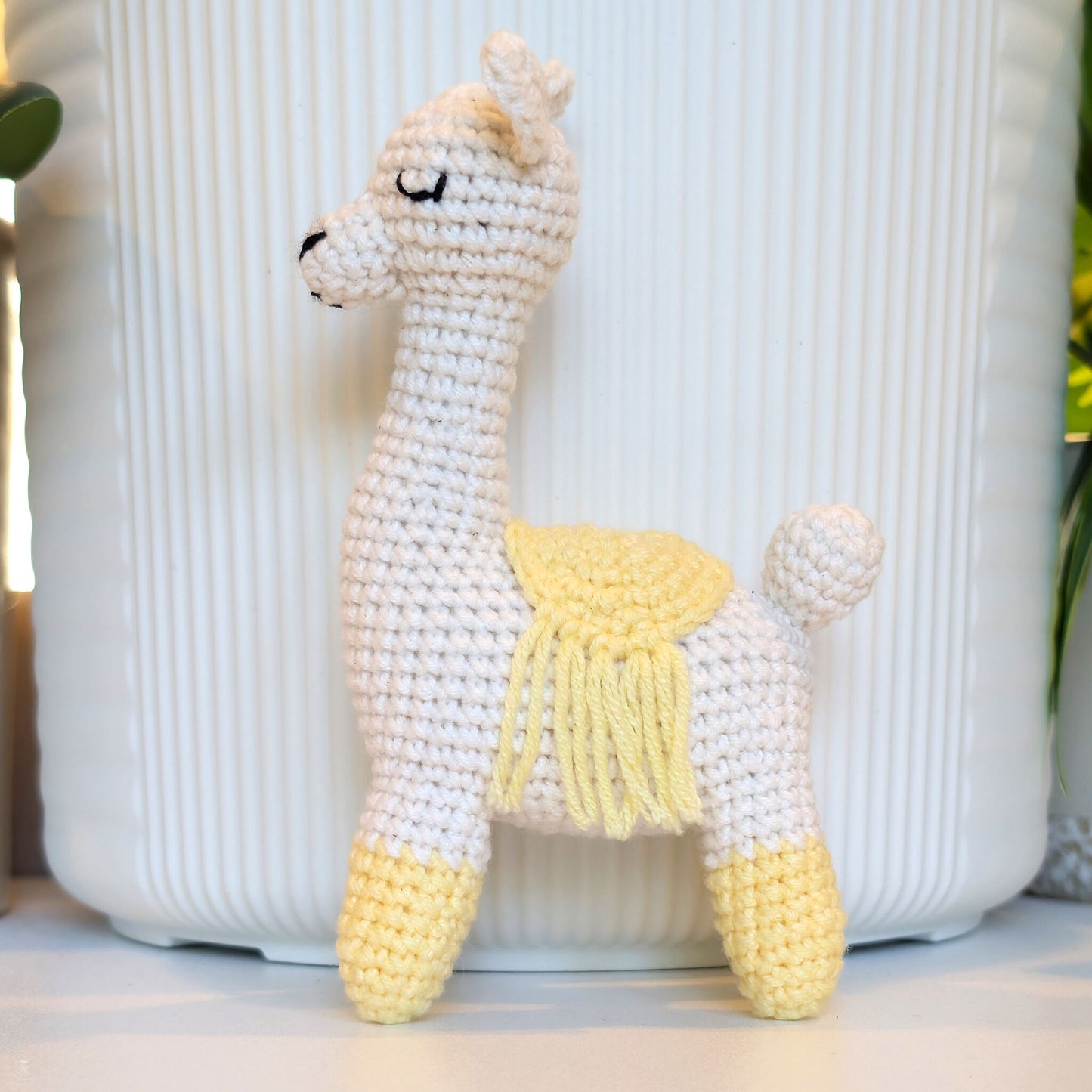 A crocheted llama toy standing in front of a white ribbed cylindrical container. The toy is primarily white with a pale yellow saddle, yellow fringe detail, and yellow booties on its feet.