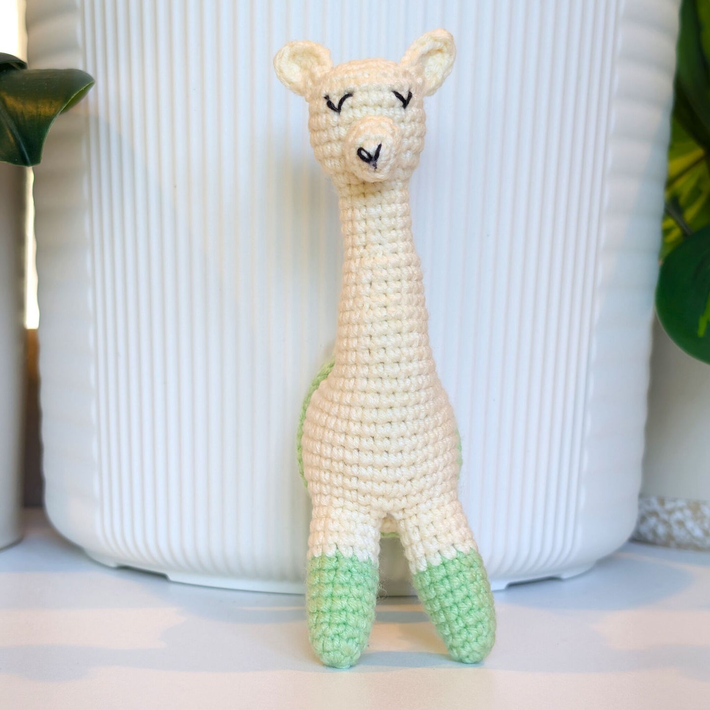 A crocheted llama toy figure with a cream-colored body, light mint green hooves, and black stitched eyes and nose, standing in front of a white ribbed ceramic pot.