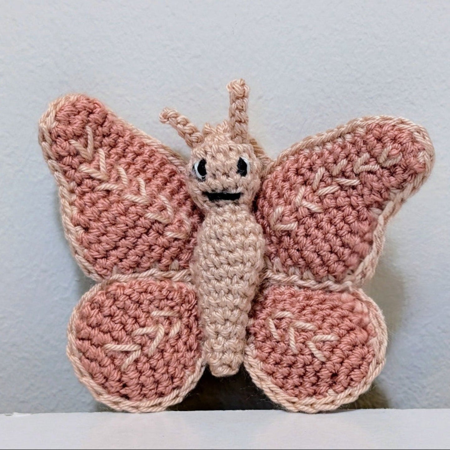 A handmade mini crocheted butterfly on string in the color dusty pink and cream, featuring a stitched smiling face, set against a light gray wall.