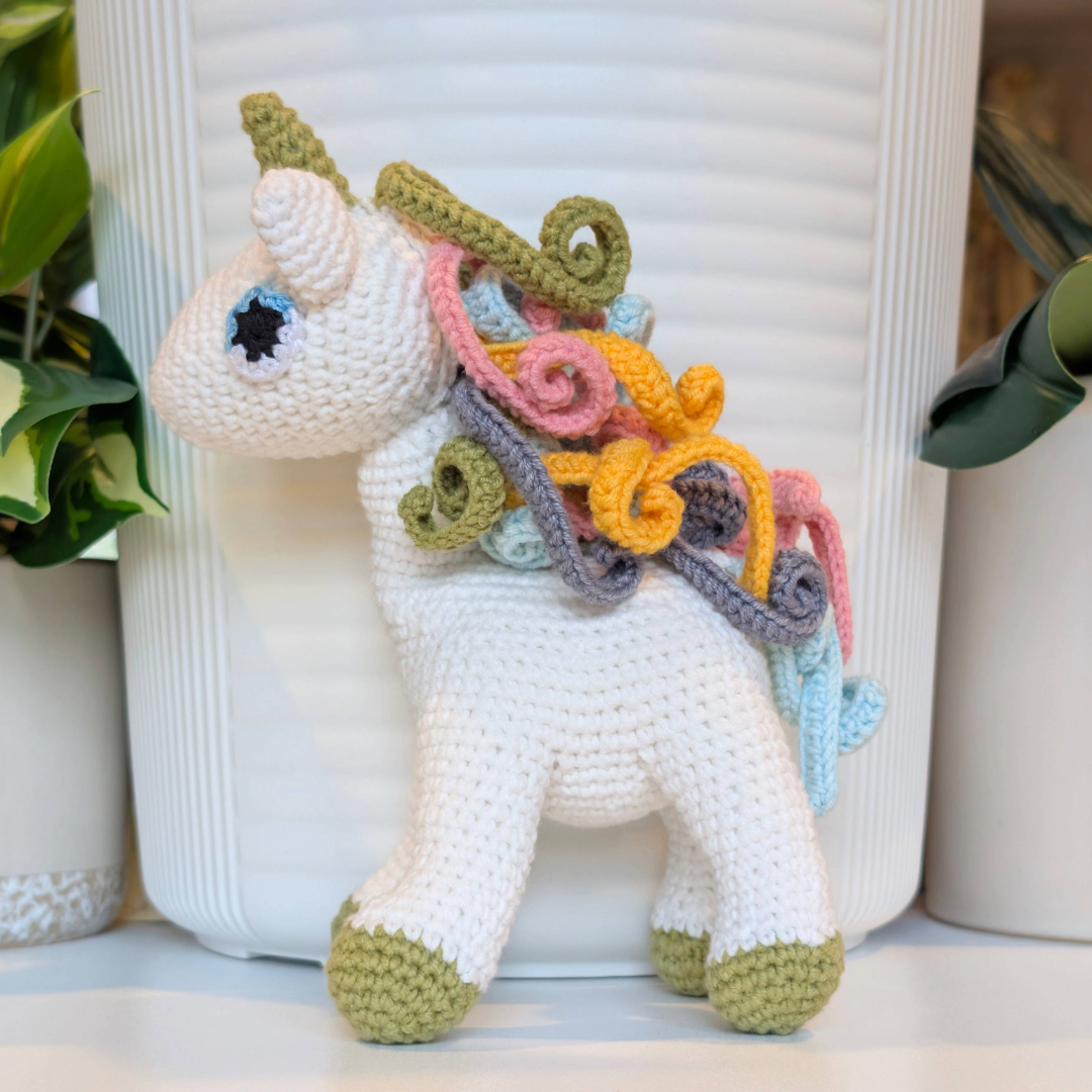Crocheted unicorn plush toy with an orange, pink, light blue and light olive green colored mane with light olive green feet.
