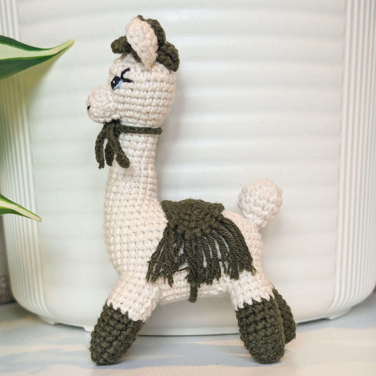 A crocheted llama toy in off-white and dark olive green yarn, standing in front of a white ribbed container with a green plant leaf visible on the left.
