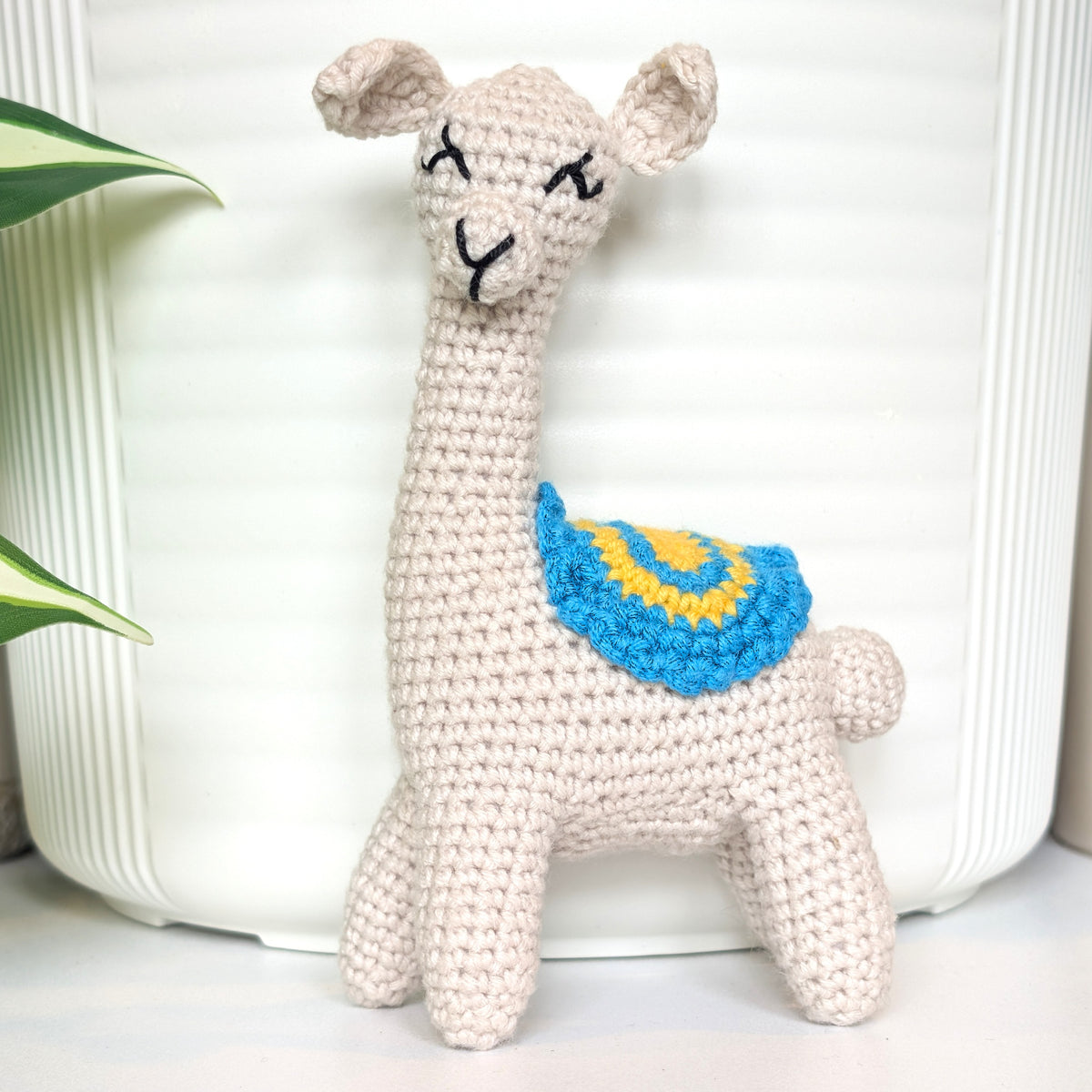 A crocheted llama toy in a light oatmeal beige color, featuring a blue and yellow spiral-patterned saddle, posed in front of a white ribbed ceramic pot with a green plant leaf visible on the left.