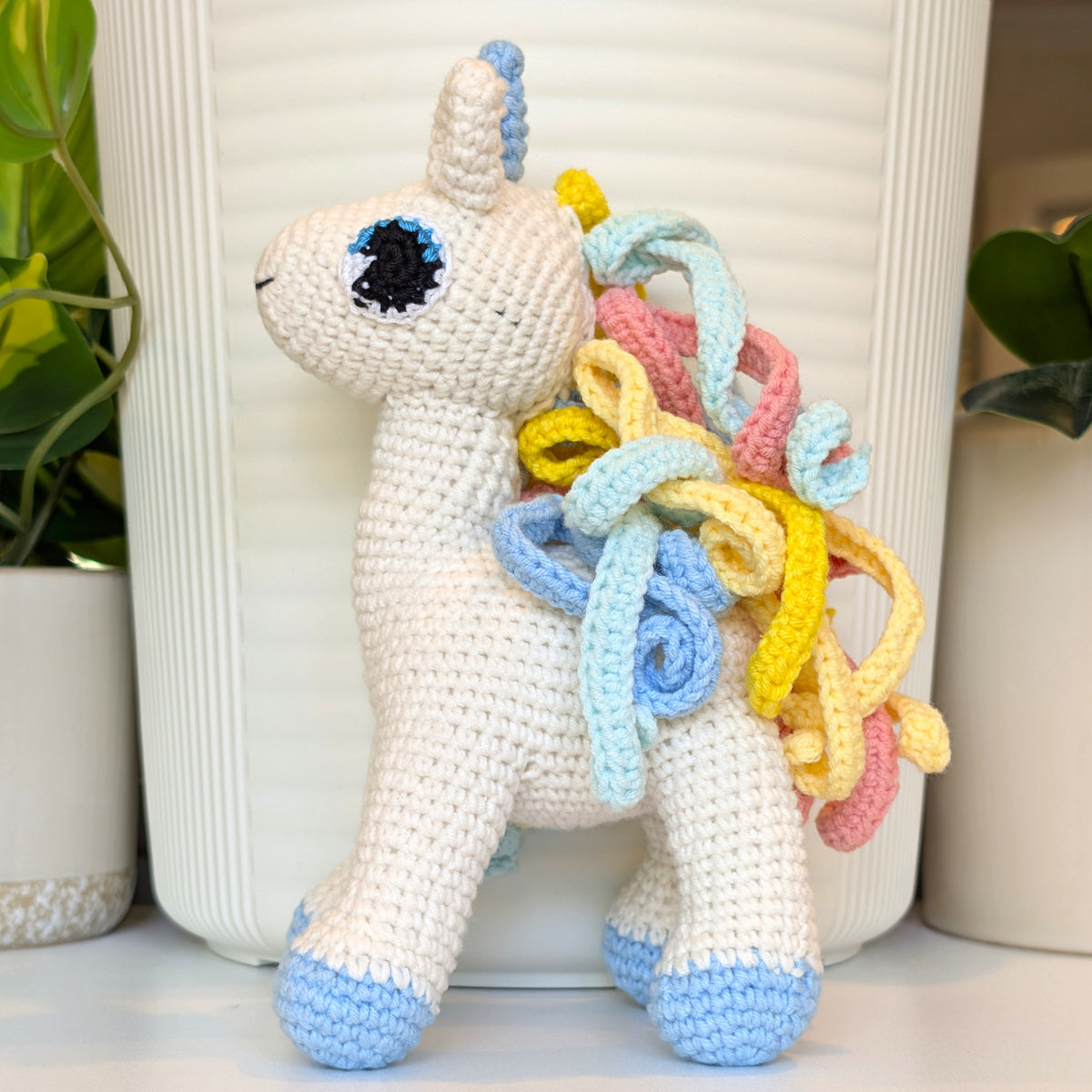 A crocheted plush unicorn toy with a cream body, light blue hooves and horn tip, and a curly multicolor mane in yellow, pink, and light blue, posed in front of a white ribbed planter with potted plants in the background.