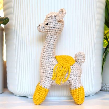 A handmade crocheted alpaca toy with a cream body, amber feet, and an amber saddle, standing in front of a white ribbed ceramic pot.