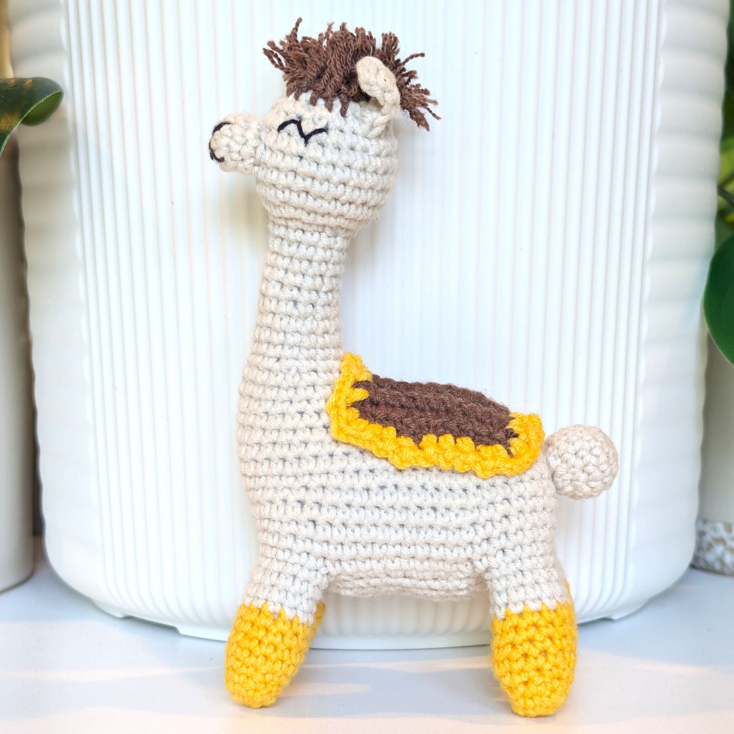 A handmade crocheted llama toy standing in front of a white ribbed ceramic pot, featuring a cream body, brown hair, a yellow and brown saddle, and yellow hooves.