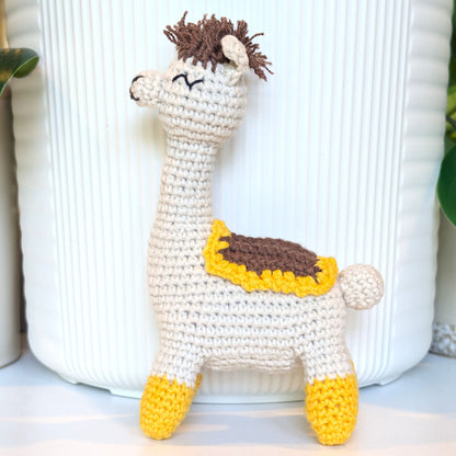 A handmade crocheted llama toy standing in front of a white ribbed ceramic pot, featuring a cream body, brown hair, a yellow and brown saddle, and yellow hooves.