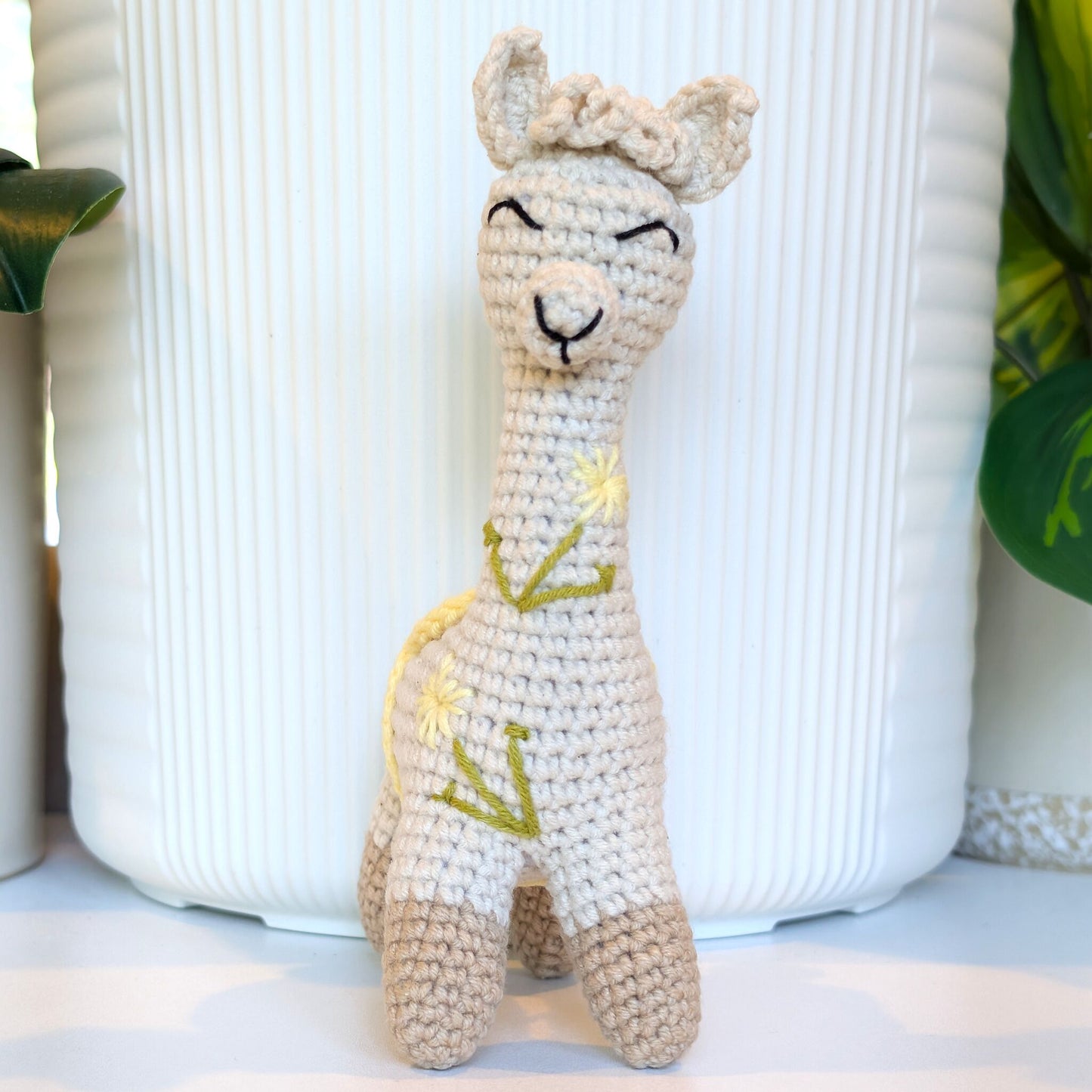 A crocheted alpaca plush toy with a cream-colored body, beige legs, and green and yellow embroidered details, standing in front of a white ribbed ceramic pot.