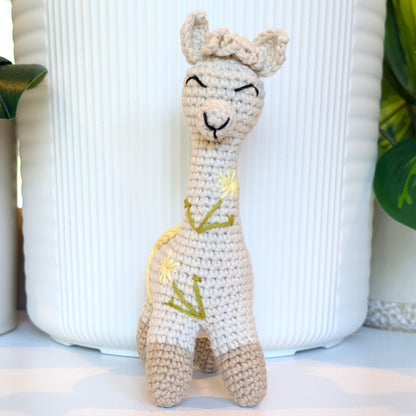 A crocheted alpaca plush toy with a cream-colored body, beige legs, and green and yellow embroidered details, standing in front of a white ribbed ceramic pot.