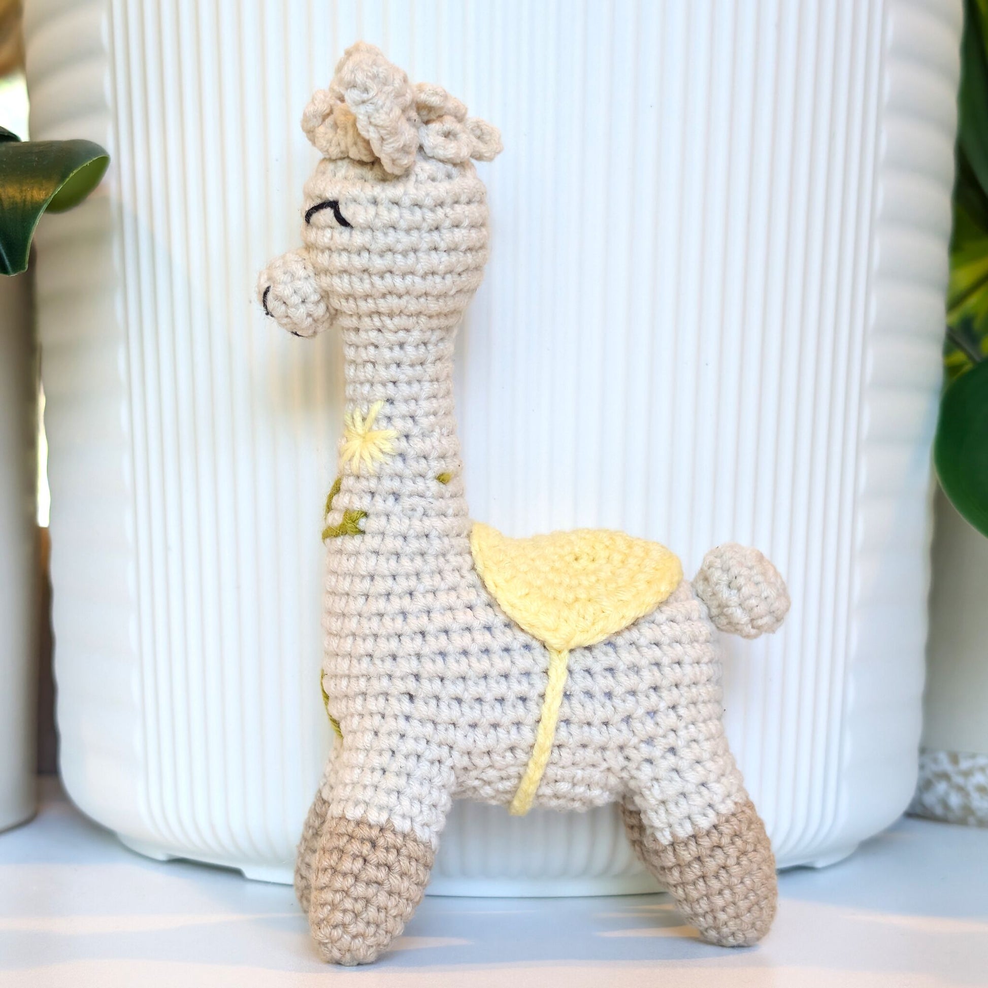 A handmade crocheted alpaca toy in cream and beige with a yellow saddle, and embroidery standing in front of a white ribbed ceramic pot.