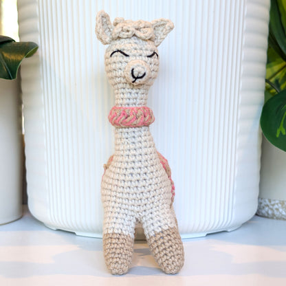 A handmade crocheted alpaca toy in beige boots with beige and pink collar, standing in front of a white ribbed ceramic pot.