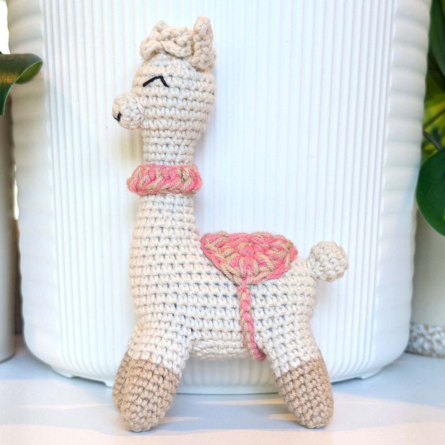 A handmade crocheted alpaca toy in beige boots with beige and pink collar, standing in front of a white ribbed ceramic pot.