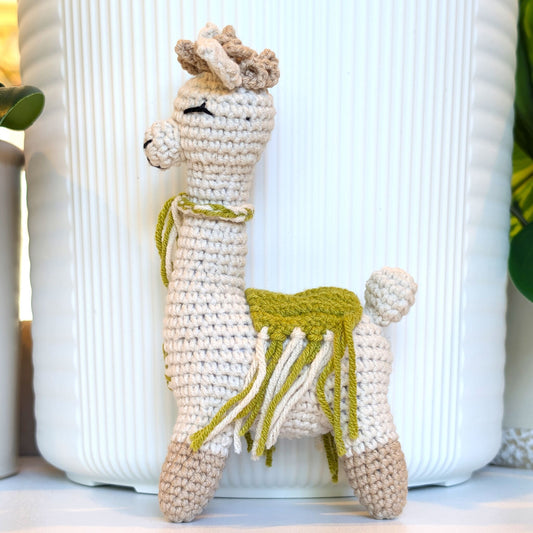 A handmade crocheted alpaca toy with a cream body, beige hooves and ears, and an pistachio green saddle and neck accessory, standing in front of a white ribbed ceramic pot.