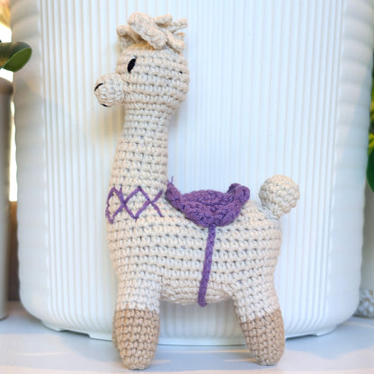 A crocheted alpaca toy with a cream-colored body, beige hooves, and a purple saddle, standing in front of a white ribbed ceramic pot.