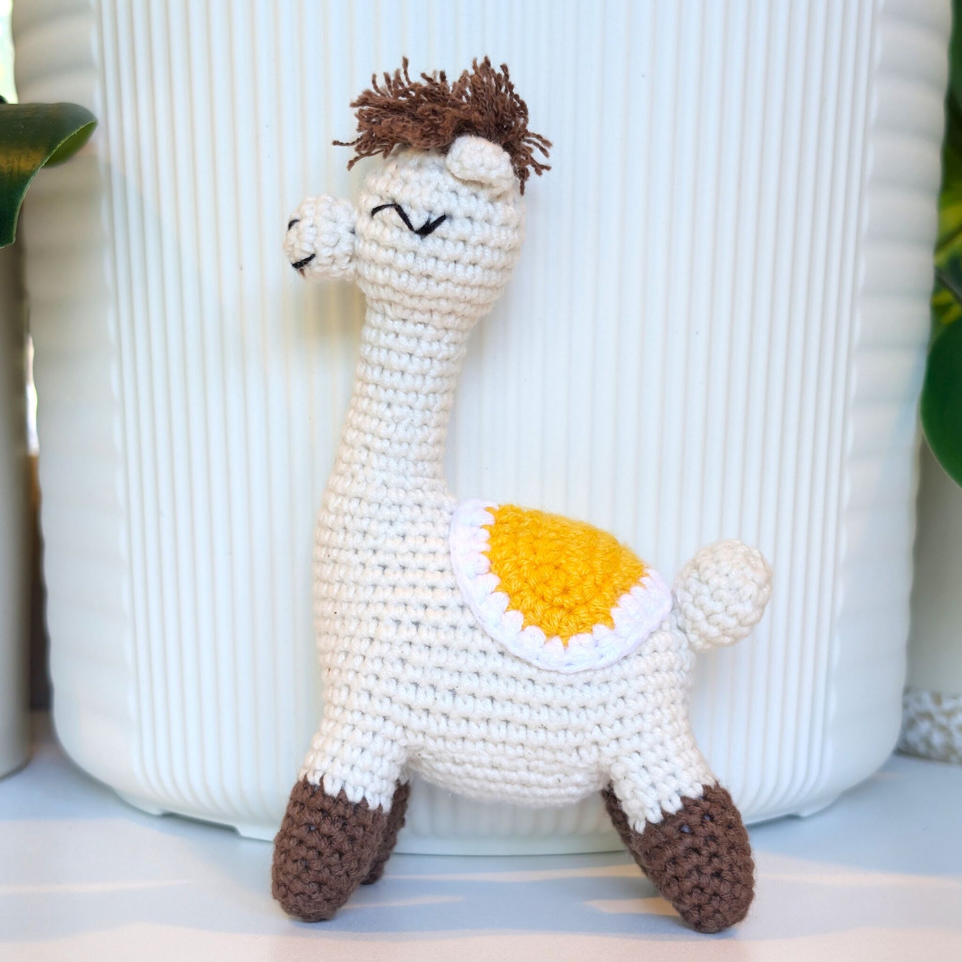 A crocheted alpaca plush toy with a cream body, brown hair, brown hooves, and a yellow-and-white saddle on its back, standing in front of a white ribbed planter.