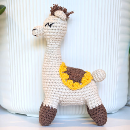 A handmade crocheted alpaca toy in cream, brown, and yellow, standing in front of a white ribbed ceramic vase.
