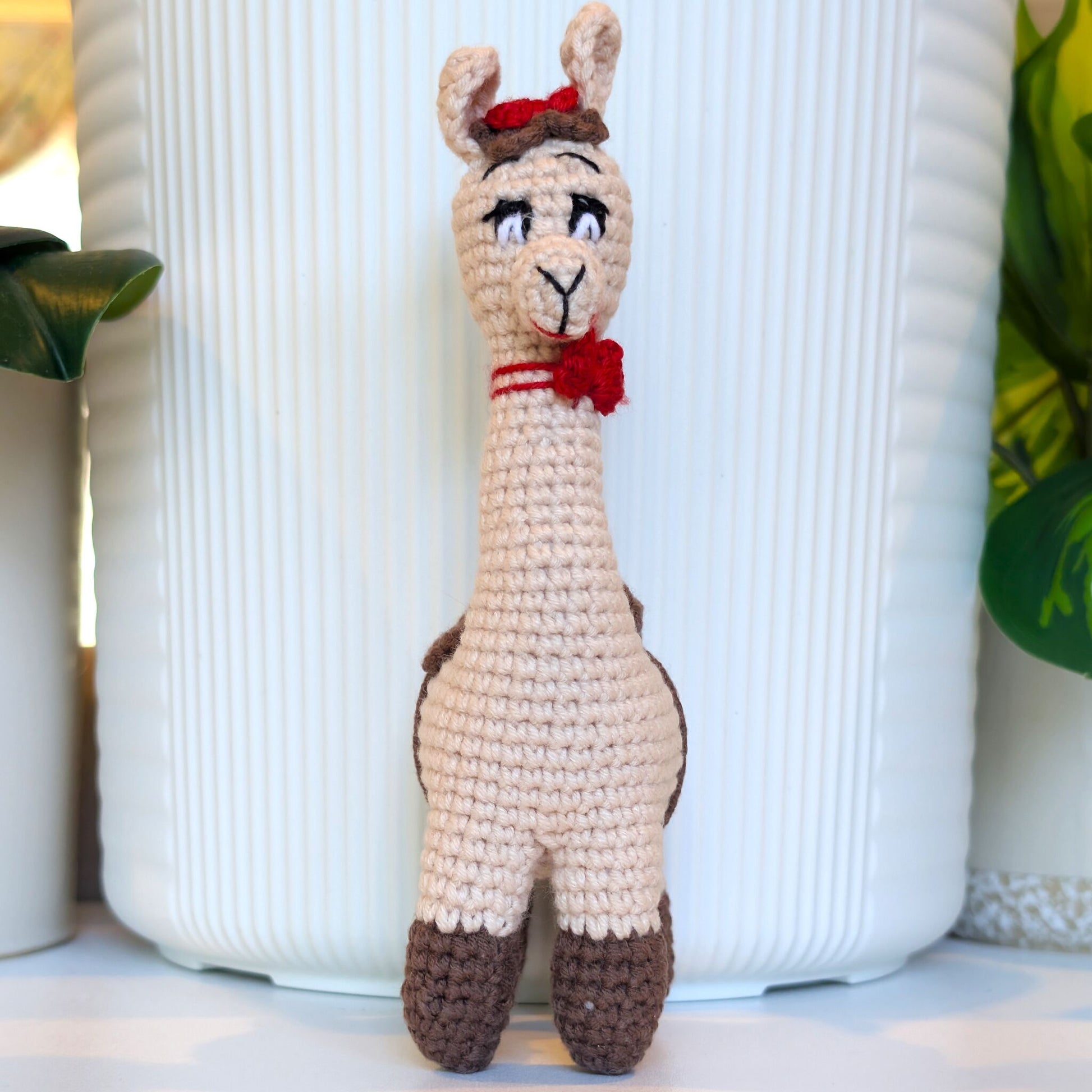 A crocheted alpaca toy with red bows standing upright in front of a white ribbed ceramic vase
