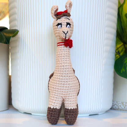 A crocheted alpaca toy with red bows standing upright in front of a white ribbed ceramic vase