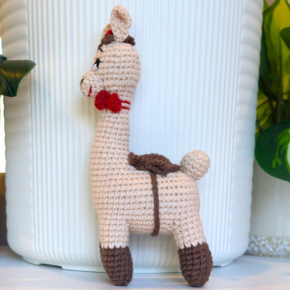 A crocheted alpaca toy toy with red bows standing in front of a white ribbed cylindrical planter