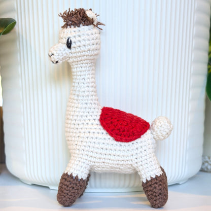 A handmade crocheted alpaca toy standing in front of a white ribbed ceramic pot. The toy is primarily off-white with brown hooves and mane, featuring a bright red saddle on its back.