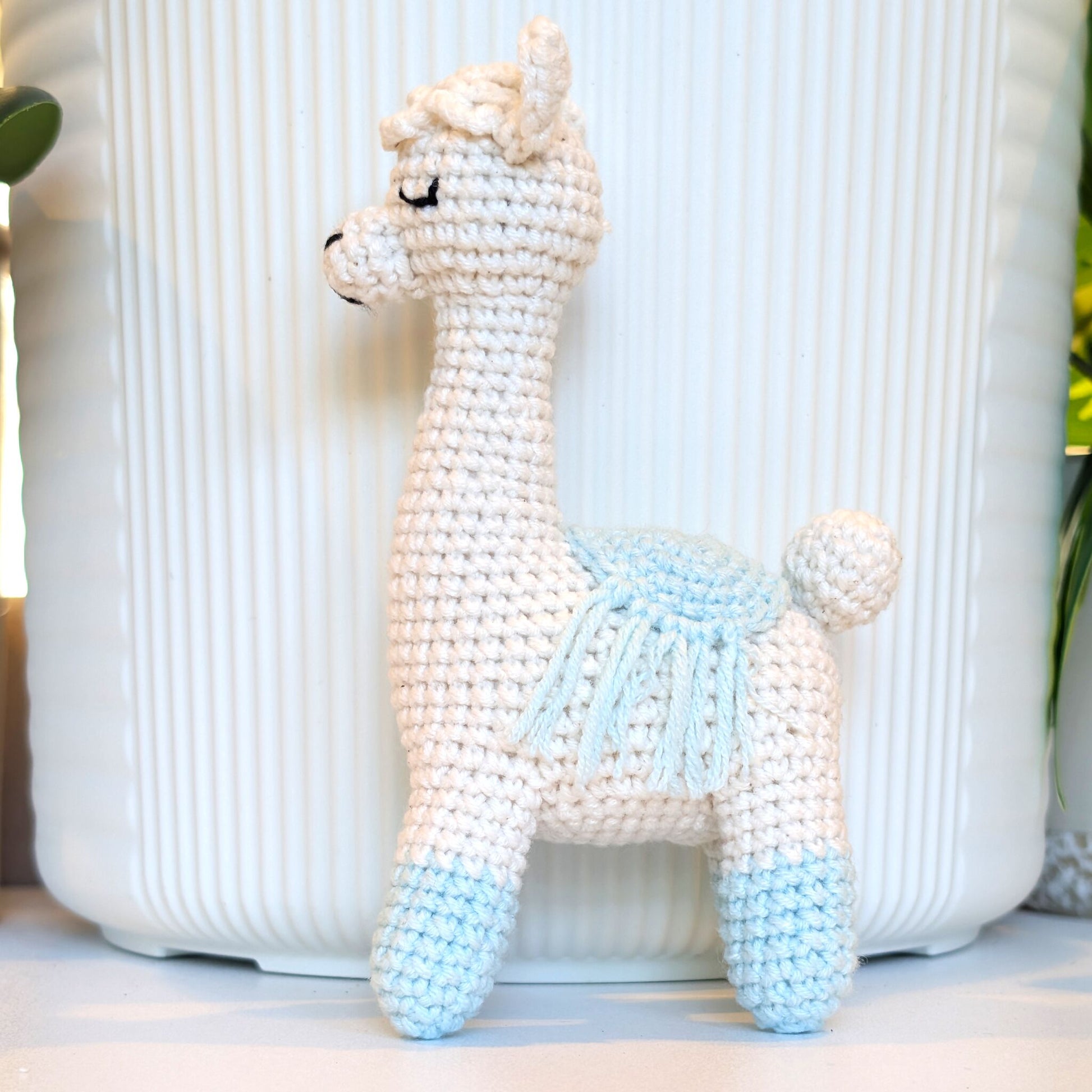 A crocheted llama plush toy in off-white with light blue or aqua accents on its legs and saddle, standing in front of a white ribbed ceramic pot.