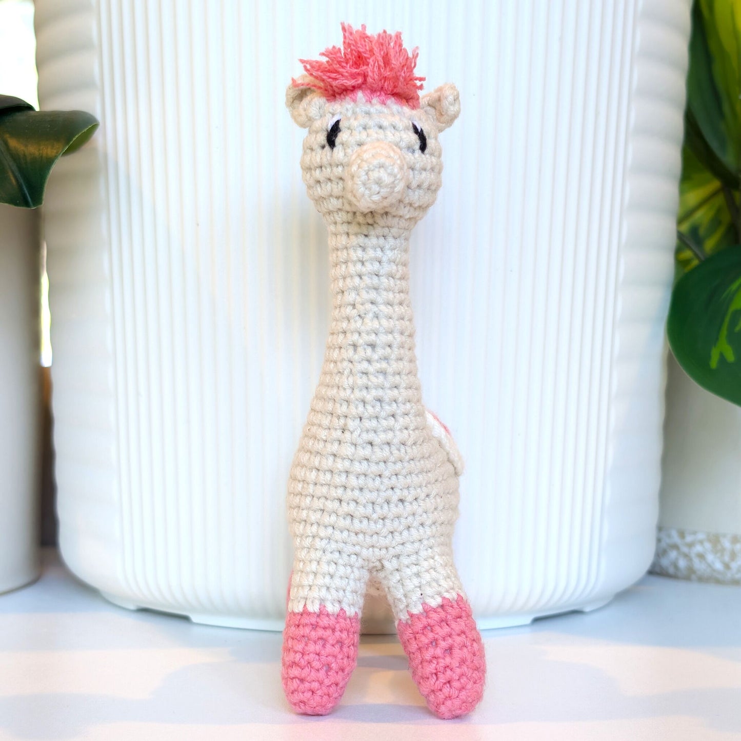 A handmade crocheted alpaca toy in cream and pink saddle and pink hair, standing in front of a white ribbed ceramic pot.