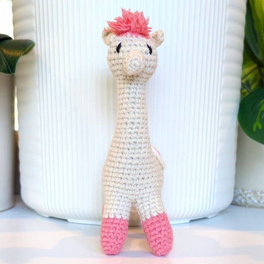 A handmade crocheted alpaca toy in cream and pink saddle and pink hair, standing in front of a white ribbed ceramic pot.