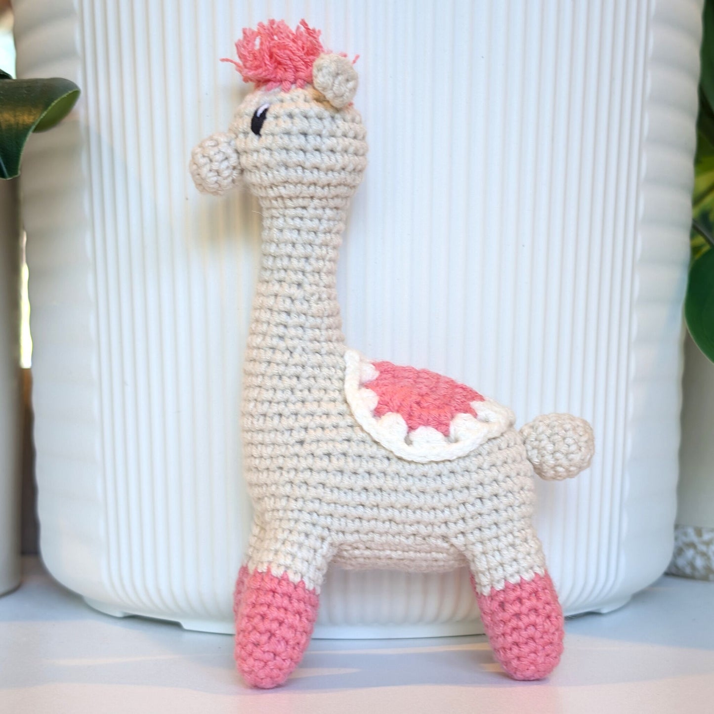 A handmade crocheted alpaca toy in cream and pink saddle and pink hair, standing in front of a white ribbed ceramic pot.