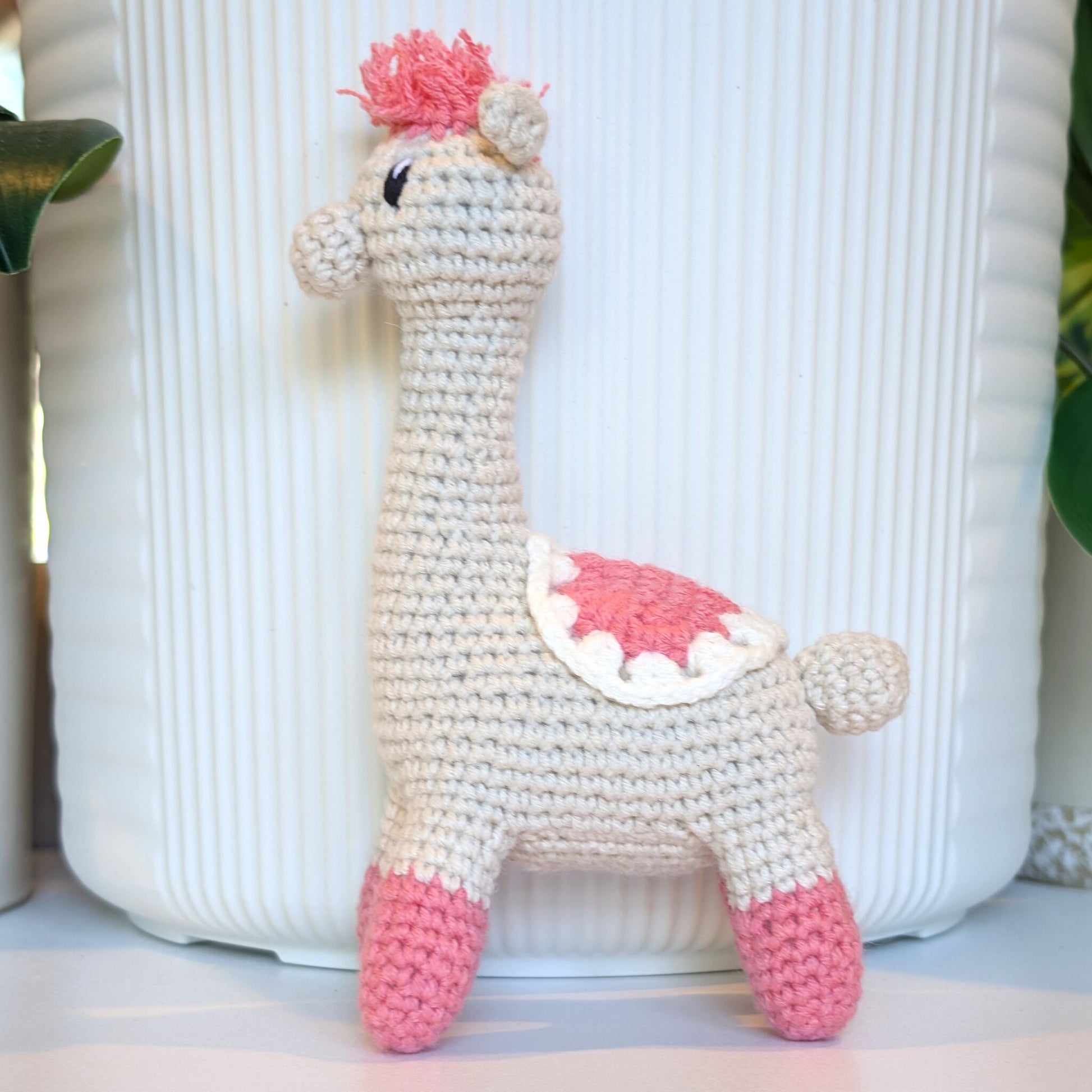 A handmade crocheted alpaca toy in cream and pink saddle and pink hair, standing in front of a white ribbed ceramic pot.