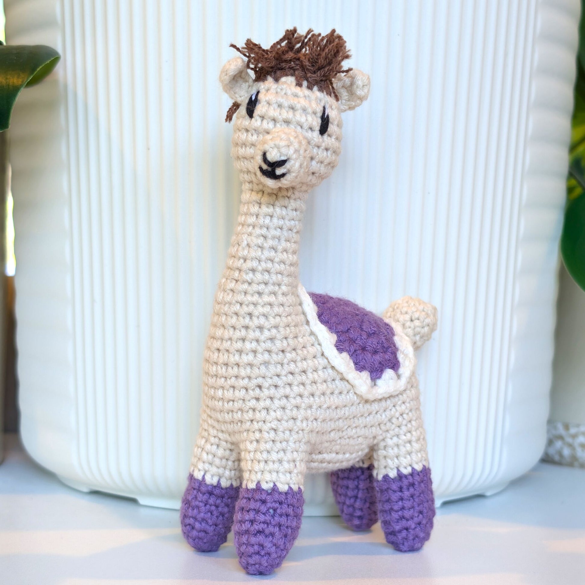 A handmade crocheted alpaca toy standing in front of a white ribbed ceramic pot. The toy features an off-white body, purple hooves and back patch, and brown yarn hair.