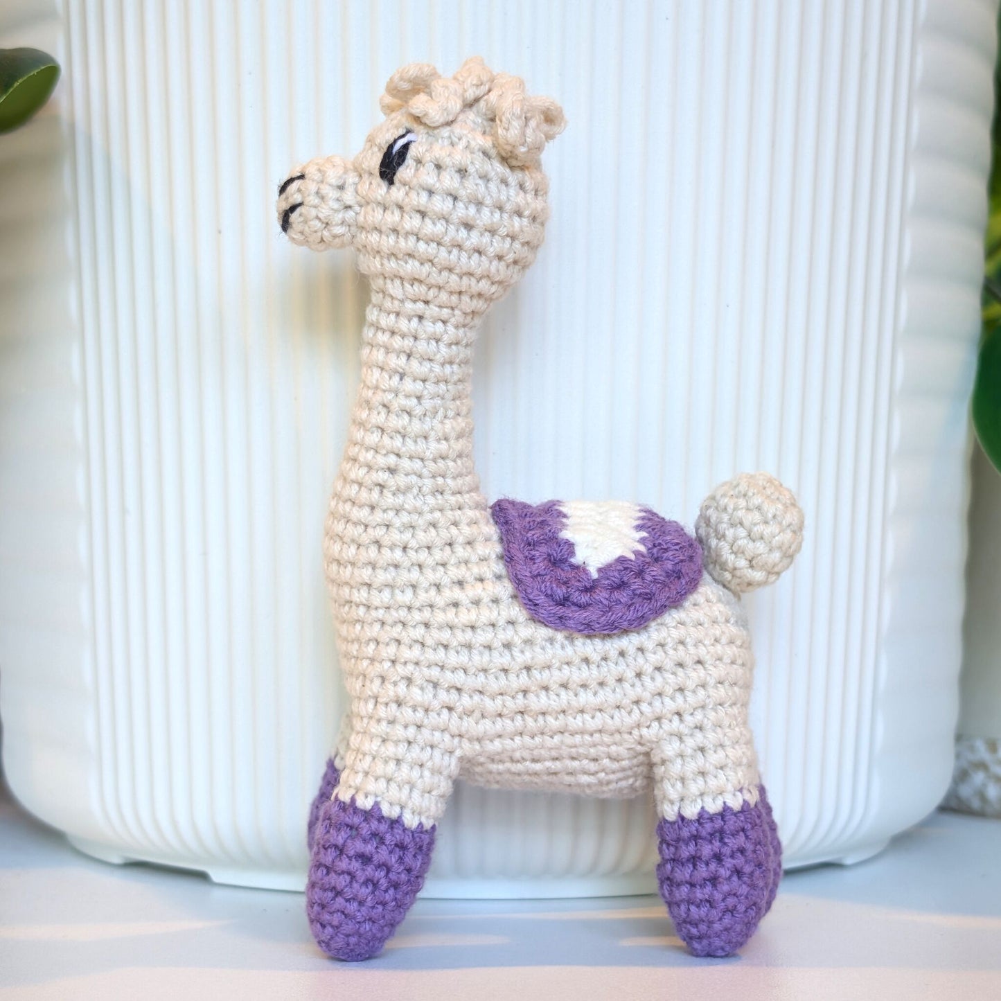 A handmade crocheted alpaca toy in cream and purple, standing in front of a white ribbed ceramic pot.