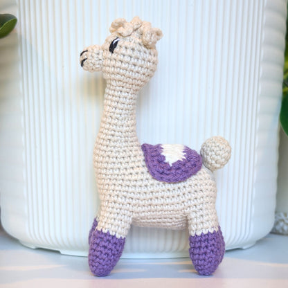 A handmade crocheted alpaca toy in cream and purple, standing in front of a white ribbed ceramic pot.