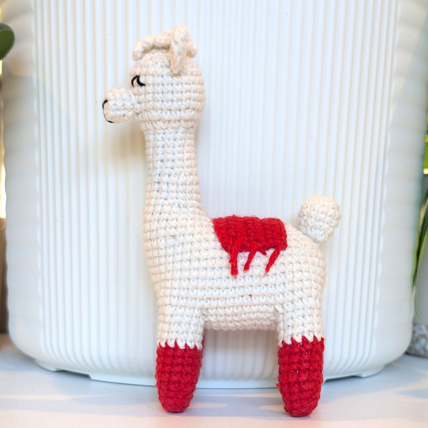 A crocheted llama toy in white with red accents on its saddle and hooves, standing in front of a white ribbed ceramic pot.